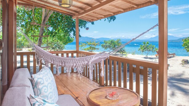 A wooden deck with a hammock, a sofa, and a round table with a bowl. - Kuda Laut Boutique Dive Resort (Siladen Island)