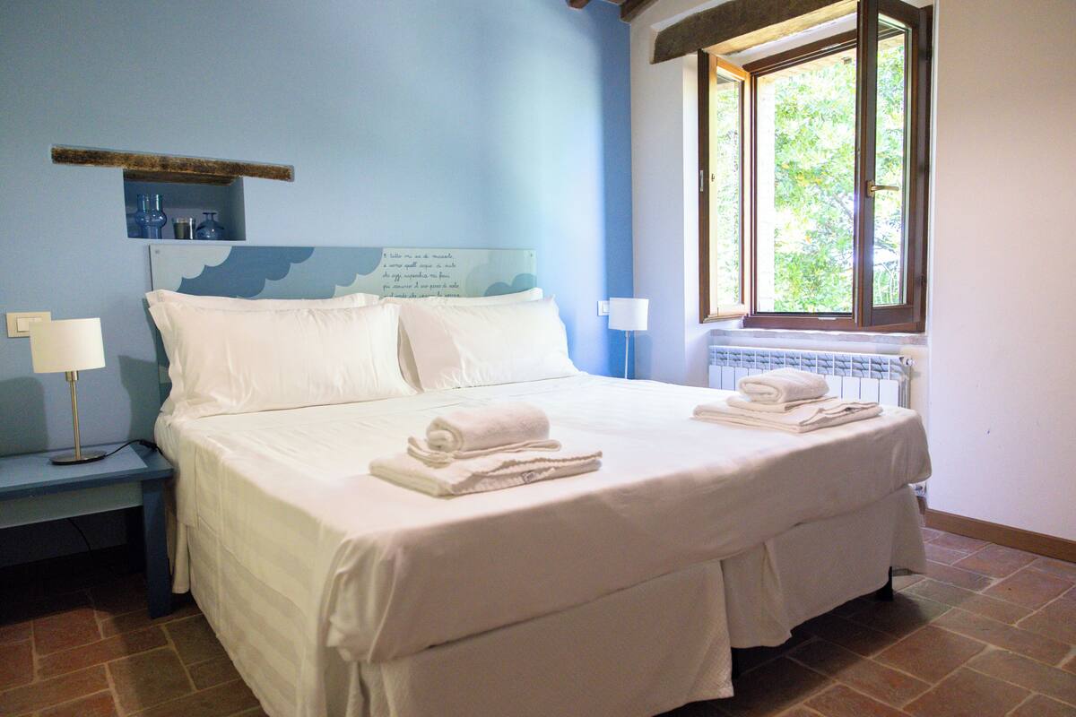 Una cama bien hecha con sábanas y toallas blancas, una mesita de noche con lámpara, y una ventana con vistas a árboles.