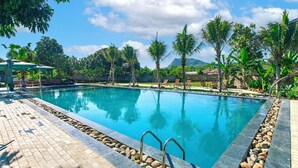 A rectangular swimming pool with a tiled deck and metal handrails, surrounded by palm trees and a stone wall. - Dong Giao Bungalow Ninh Binh (Trung Son)