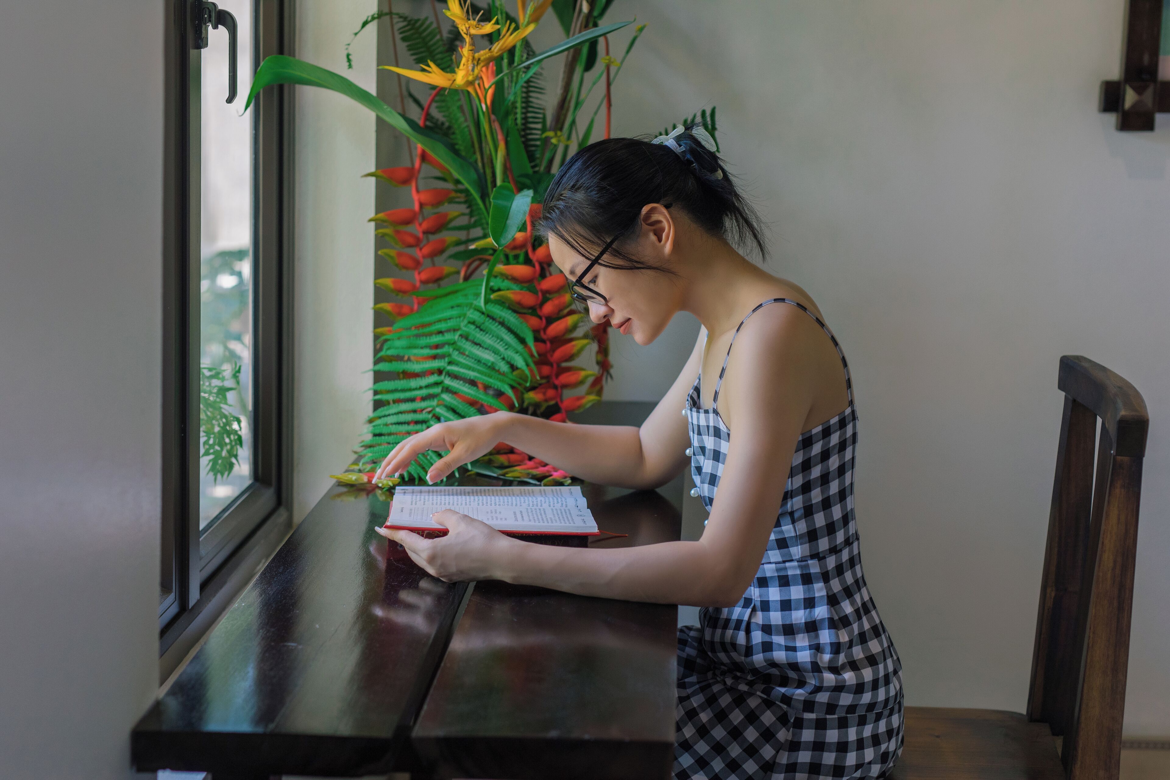 A person sitting at a wooden table with a book, a window with a view of greenery, and a large plant with red flowers in the background.