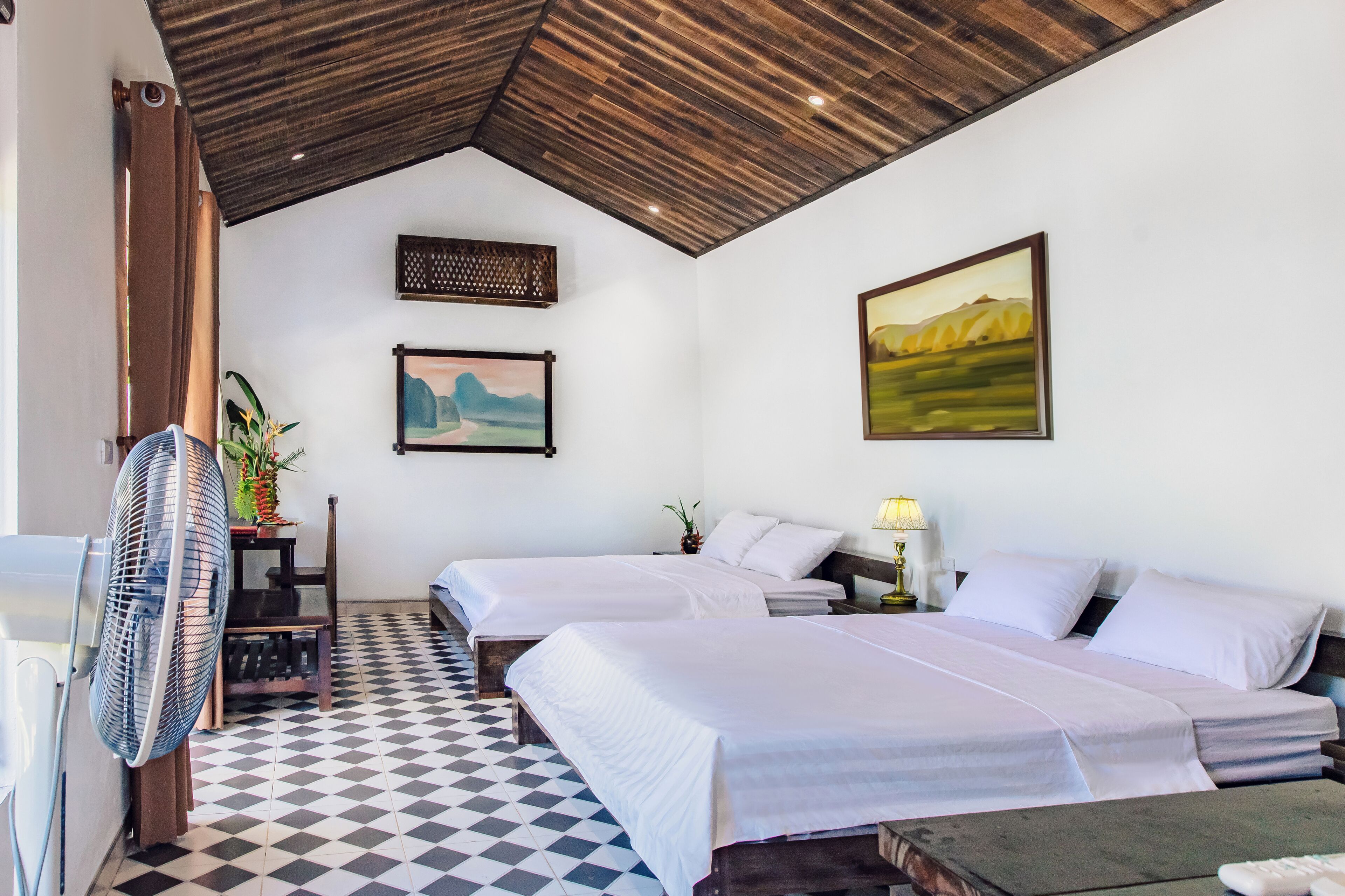 A hotel room with two beds, a checkered floor, a fan, and artwork on the walls.