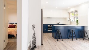 A modern kitchen with a central island, bar stools, and a view into a bedroom with a bed and a red pillow. - Brewers Haus ~ Willunga - A Newly Renovated Property In the Heart Of Willunga (Willunga)