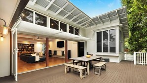 A spacious deck with a wooden table and benches, a large window, and a view of the interior living area. - Cavvanbah Beach Cottage, steps from the beach (Byron Bay)