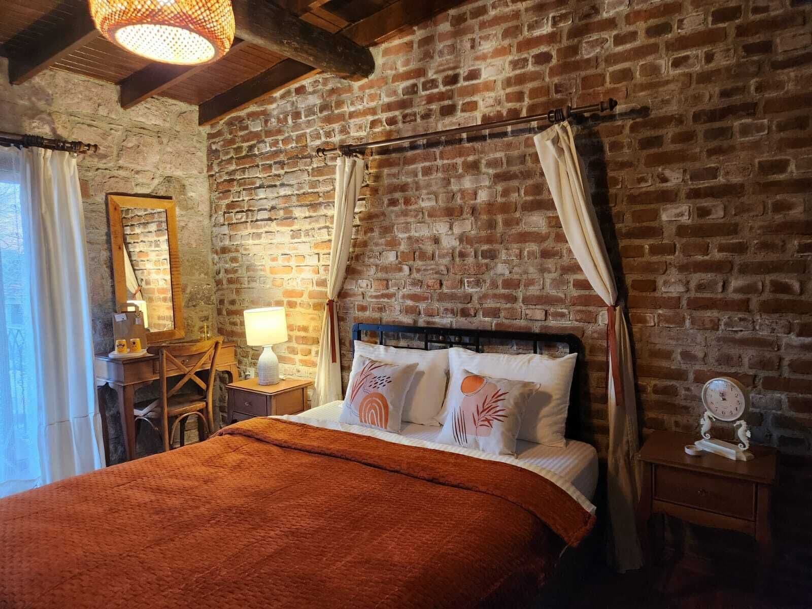 A rustic room with exposed brick walls, a wooden bed with a red blanket, a desk with a lamp, and a mirror on the wall.