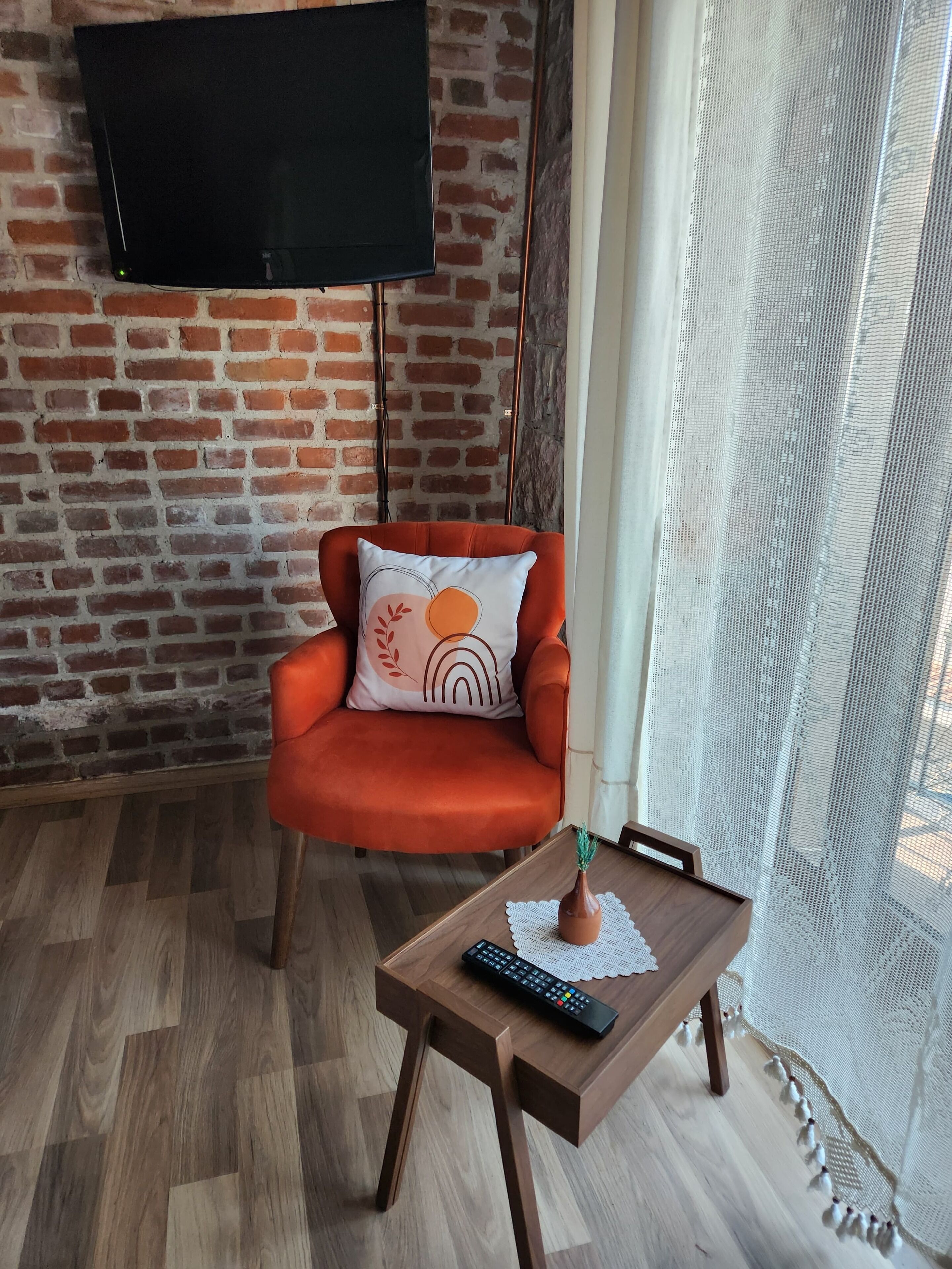 Een woonkamer met een bakstenen muur, een televisie bovenop een rode fauteuil met een decoratief kussen, een houten salontafel met een afstandsbediening, en een kantelaar.