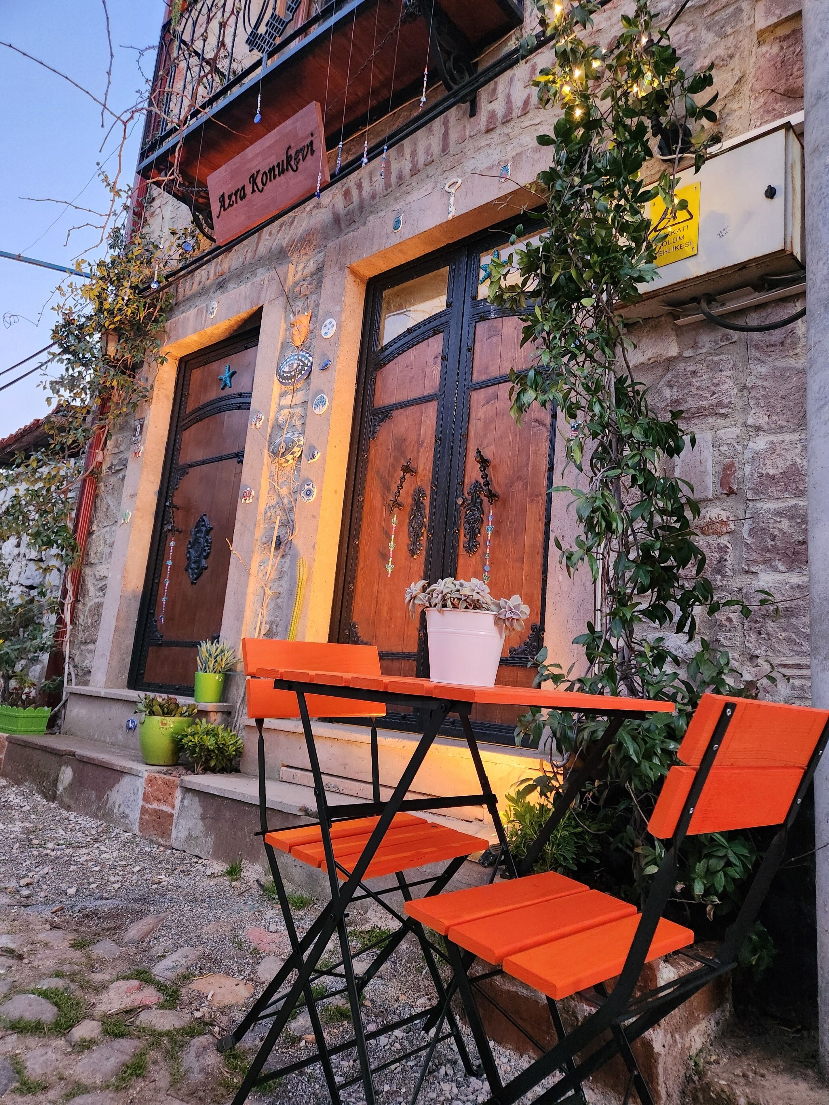 A rustic building with a wooden door and windows, outdoor seating with orange chairs and tables, and decorative elements like plants and wall art.