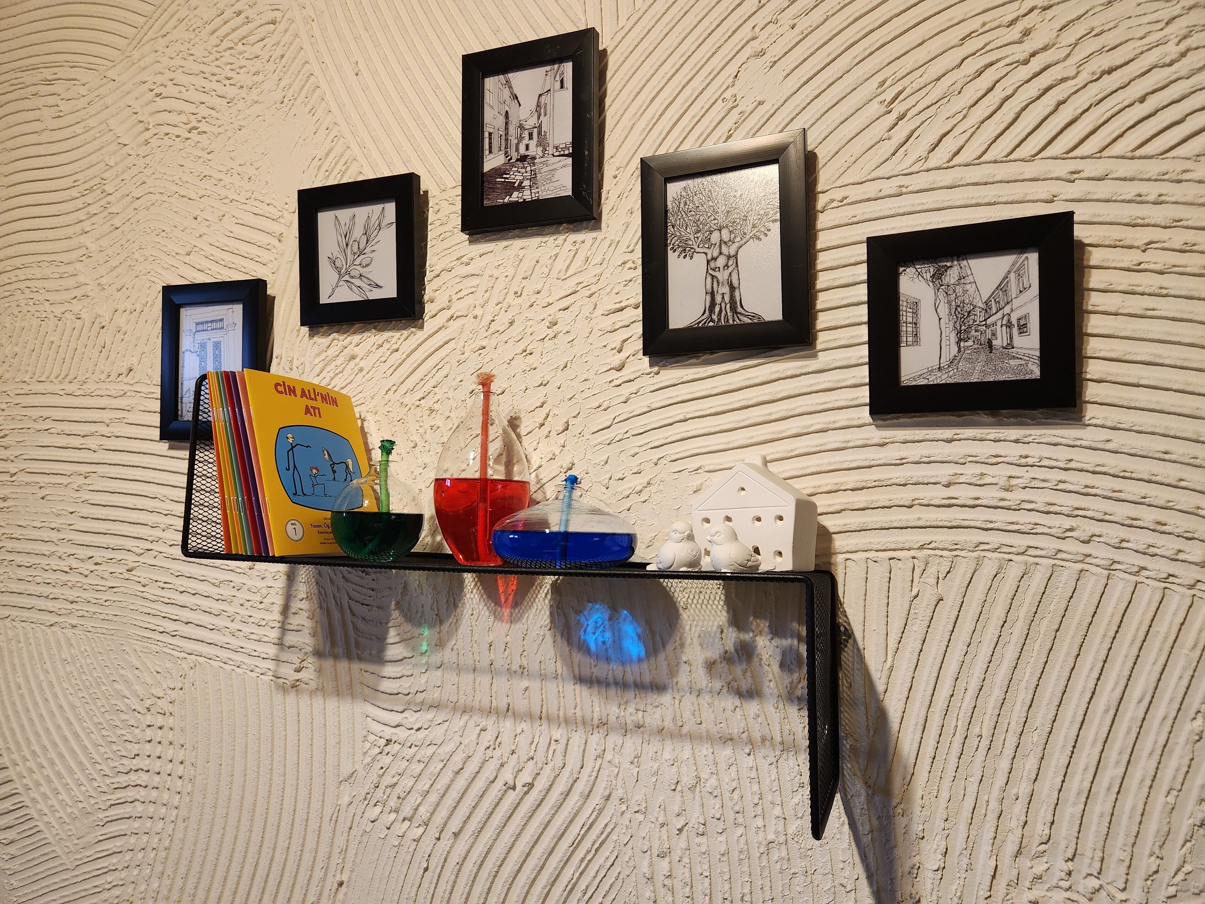 A wall with textured plaster and four framed black-and-white drawings. A shelf holds a colorful book, a green bowl, a red liquid container, and a blue bowl.
