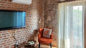 Quarto com parede de tijolos, televisão de tela plana na parede, teto de madeira, luminária de vime, poltrona acolchoada, mesa pequena com uma xícara e janela com cortinas leves.