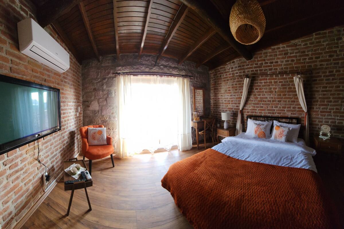 Ein Schlafzimmer mit Backsteinwand, Holzboden, einem Bett mit orangefarbener Decke, einem Fernseher, einem Sessel und einem Fenster mit Vorhängen.