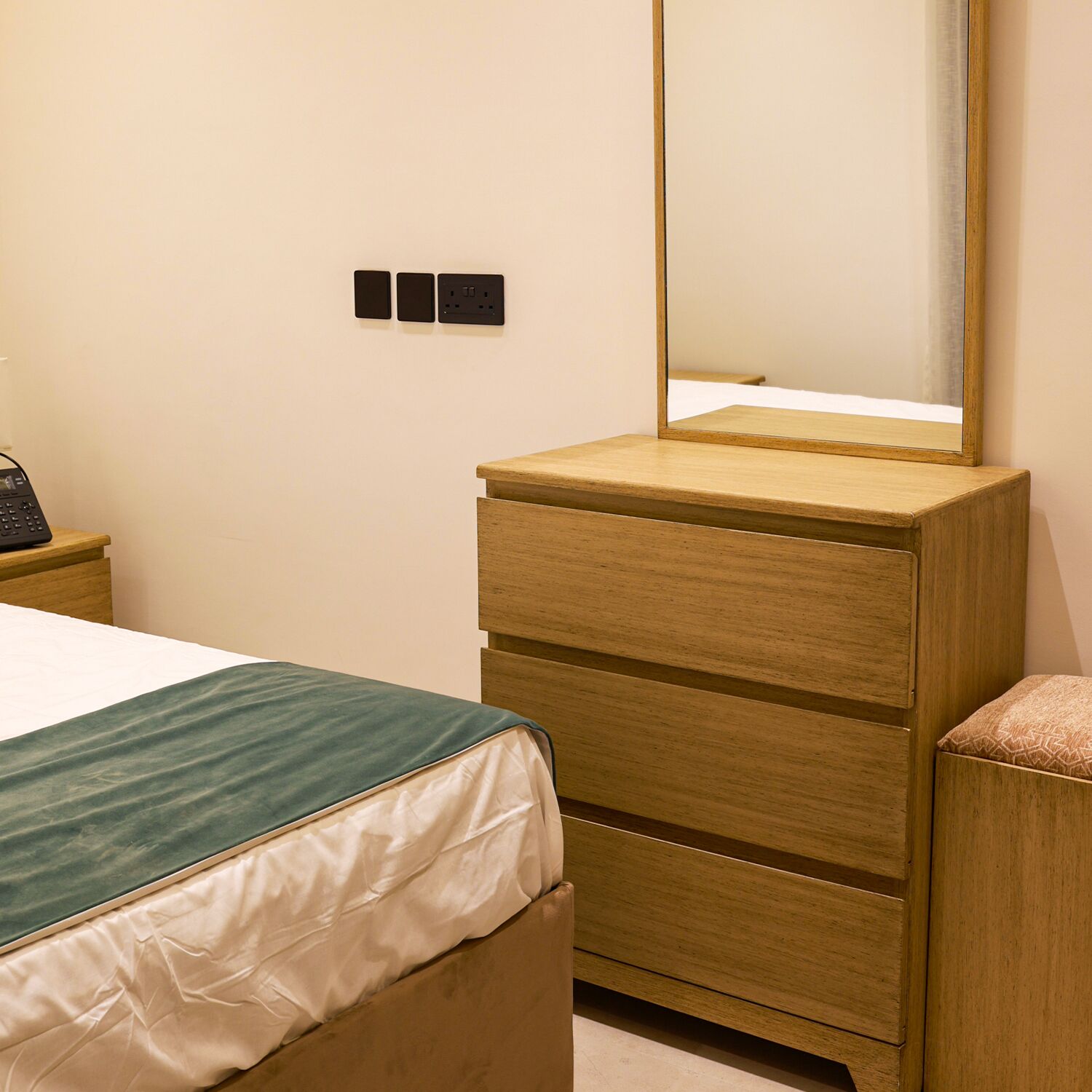 A bedroom with a bed, a wooden dresser with a mirror, and a bedside table with a phone.