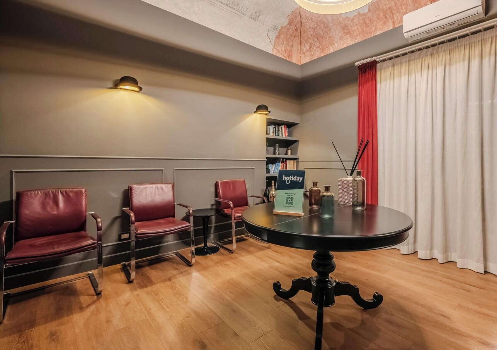 Quarto com mesa redonda, três cadeiras de couro, uma estante e uma cortina vermelha.