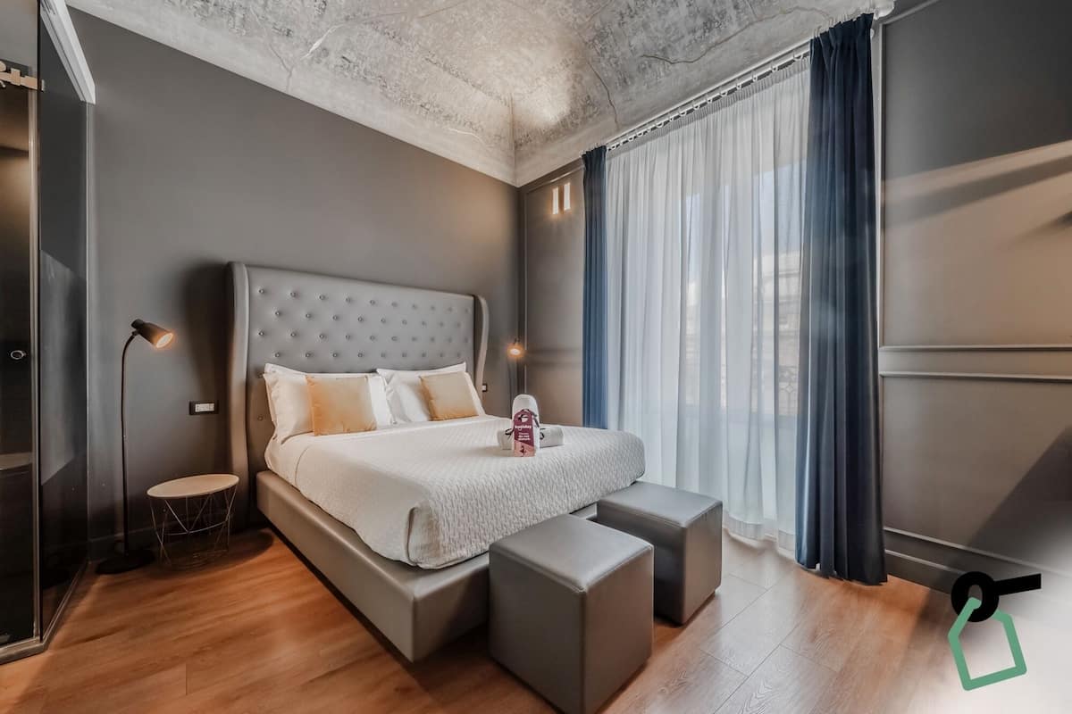A modern bedroom with a large bed, bedside tables, a floor lamp, and a view of the city through sheer curtains.