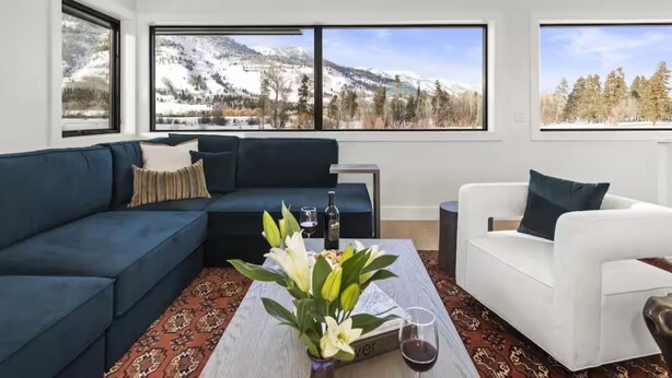 A modern living room with a dark blue sofa, a white armchair, and a coffee table with a vase of flowers and a glass of wine. Large windows offer a view of a snowy mountain landscape.