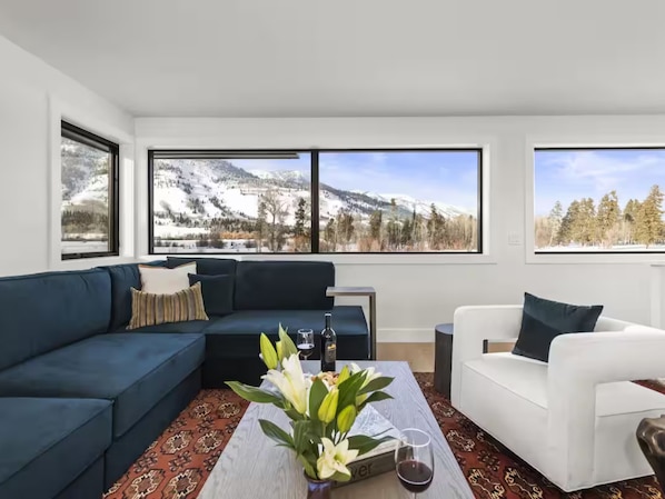 Una sala de estar moderna con un sofá azul oscuro, un sillón blanco y una mesa de centro con un jarrón de flores y una copa de vino. Amplias ventanas ofrecen vistas a un paisaje montañoso nevado.