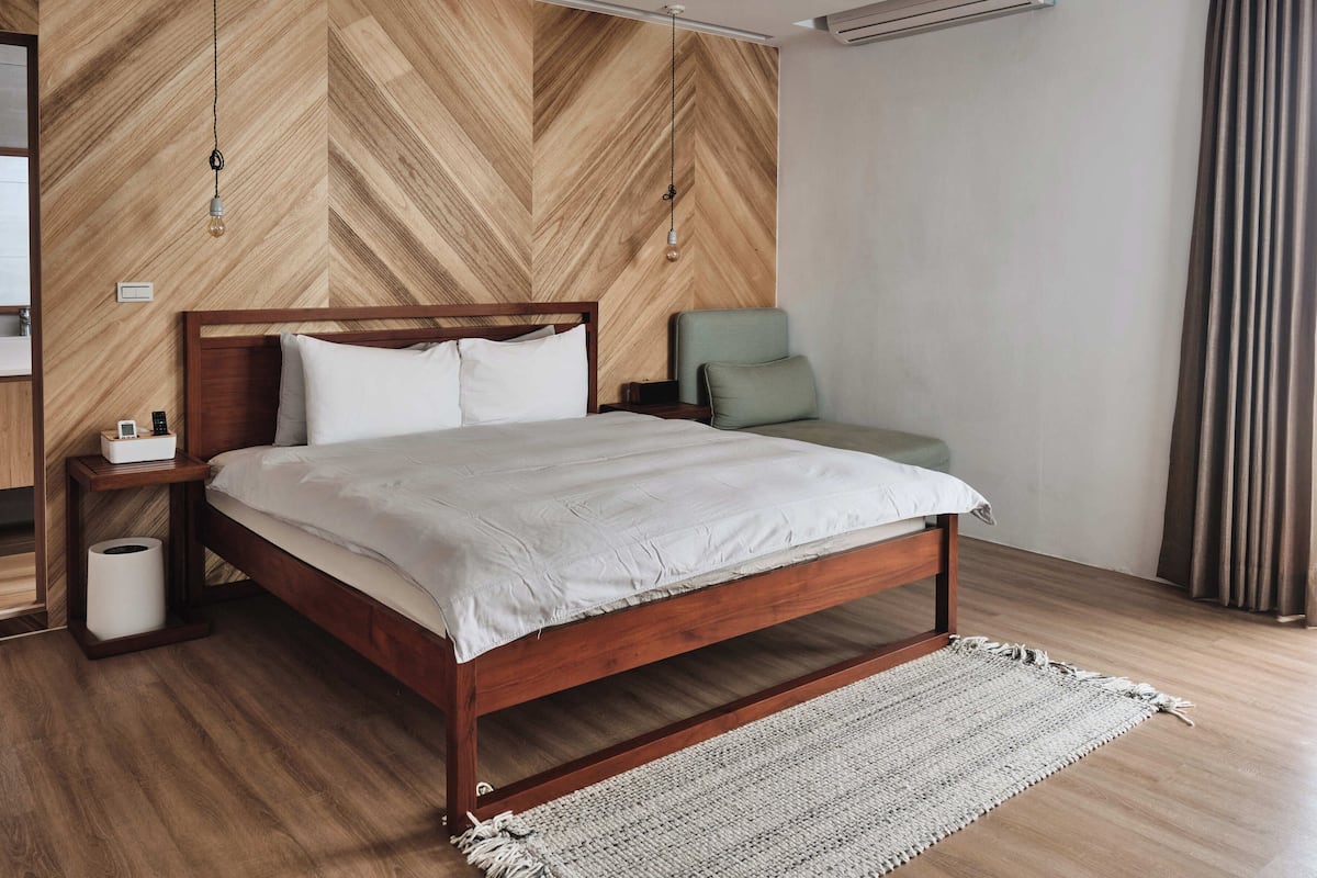 A modern bedroom with a wooden herringbone-patterned wall, a wooden bed frame, a green chair, and a white bedside table with a lamp.