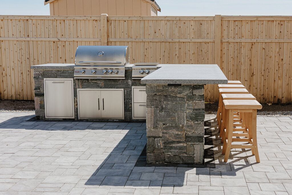 An outdoor kitchen with a stone island, a grill, and wooden stools.
