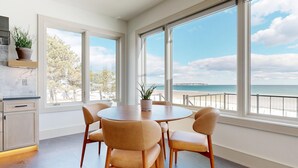 Un comedor con una mesa redonda y cuatro sillas, ventanas amplias con vista a la playa, una planta en maceta en el alféizar de la ventana y un salpicadero de mármol en la cocina.