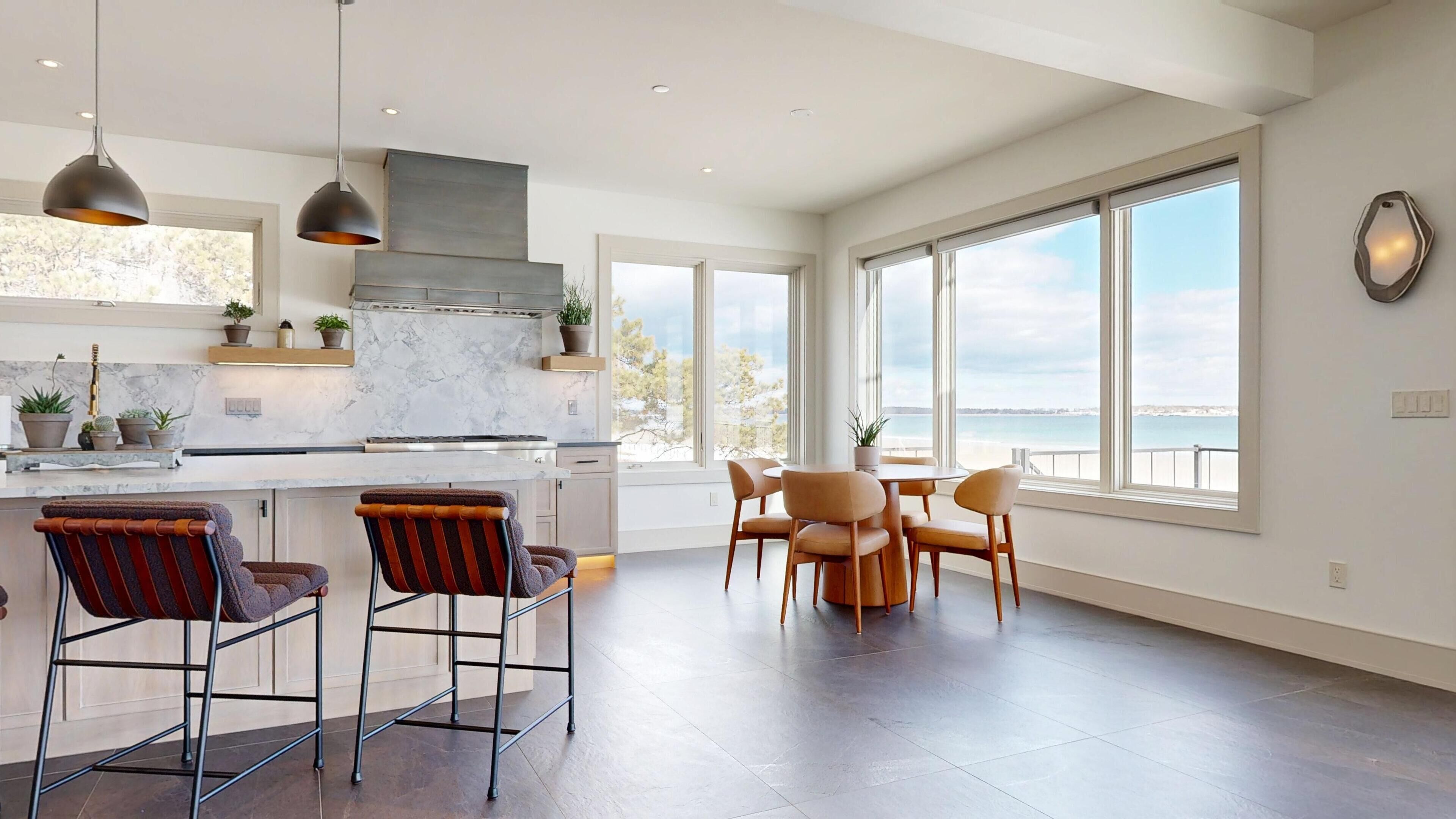 A modern kitchen with a large island, bar stools, and a dining area with a view of the sea.