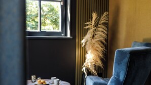 Une pièce moderne avec un fauteuil bleu, une petite table ronde sur laquelle se trouve une assiette de pâtisseries, une grande plante et une fenêtre avec des stores.