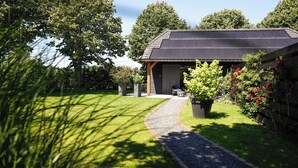 A modern house with a solar panel roof, a gravel pathway leading to the entrance, and a garden with potted plants and red flowers. - Atmospheric luxury holiday home –  spacious garden – 5 minutes from the forest. (Wilbertoord)