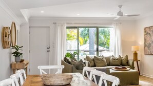 Un salon lumineux avec une table à manger en bois, des chaises blanches, un canapé avec des coussins verts, un ventilateur de plafond et une grande fenêtre avec des rideaux.