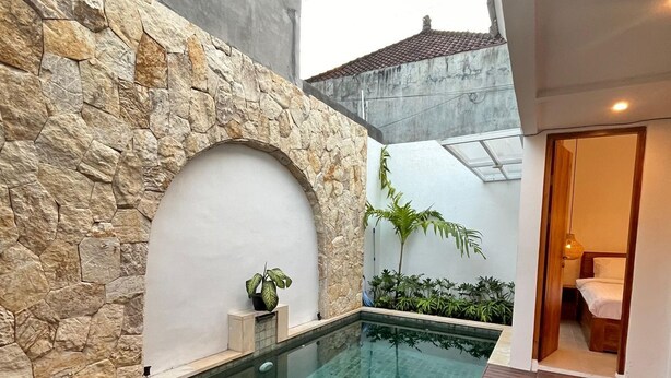 Ein Schwimmbecken mit Steinmauer und Holzterrasse, umgeben von Pflanzen und einem kleinen Garten.