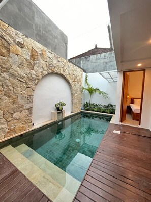 A swimming pool with a stone wall and wooden deck, surrounded by plants and a small garden. - Della MIA (Kerobokan)