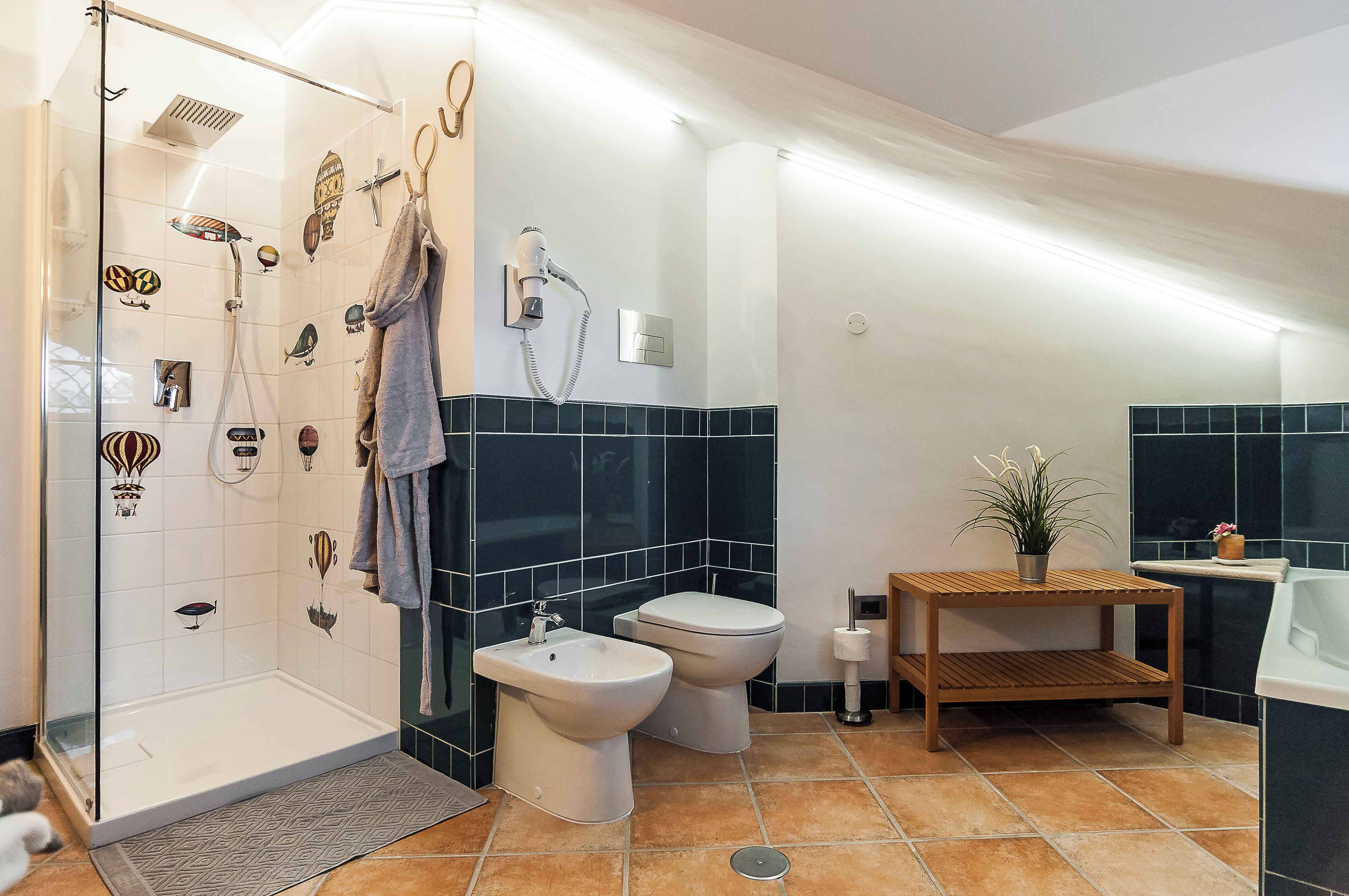 Un bagno moderno con box doccia in vetro, due bidet, un water e un piccolo tavolo in legno con una pianta.