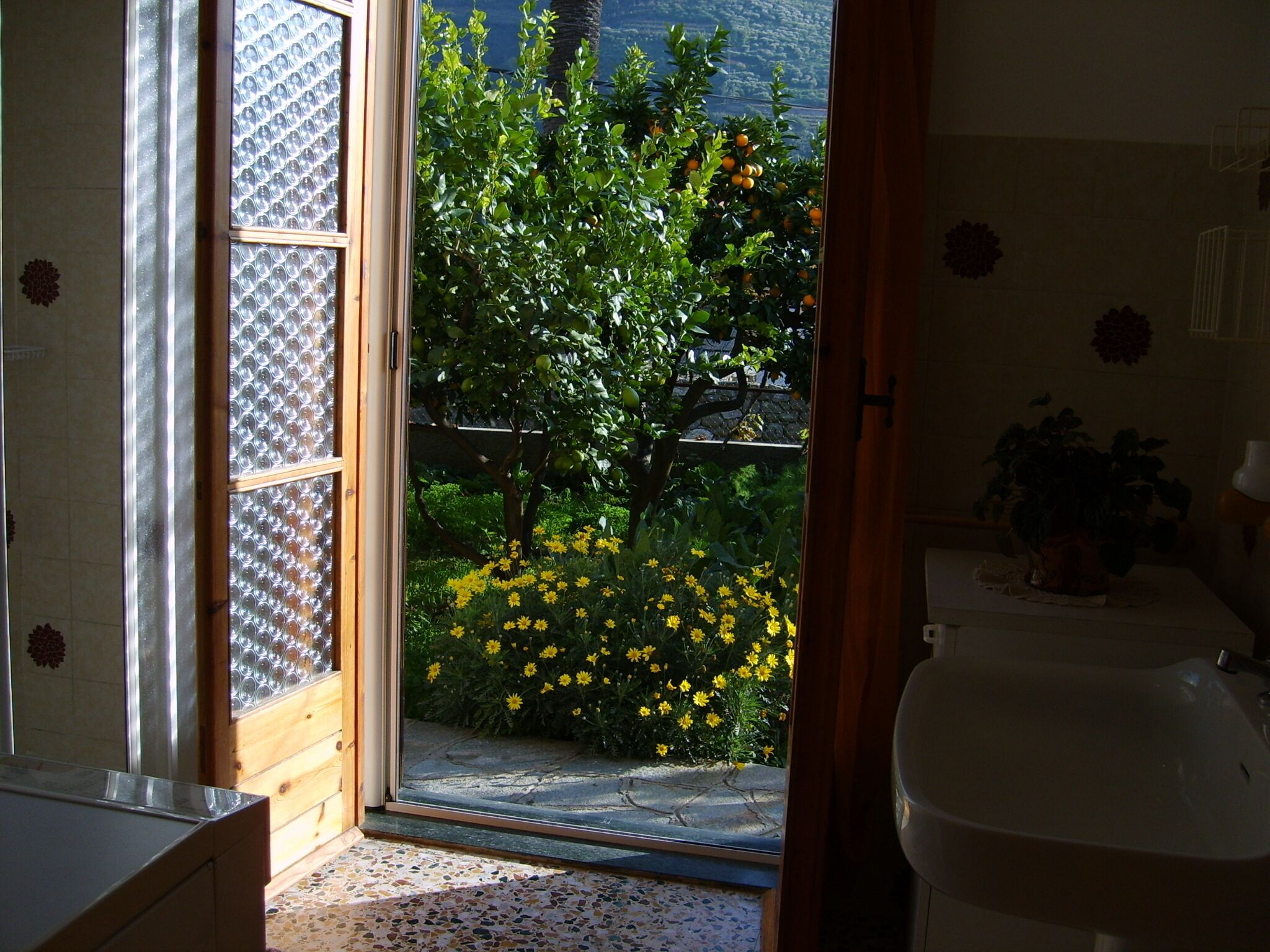 Ein Badezimmer mit Waschbecken, Spiegel und Fenster mit Blick auf einen Garten mit gelben Blumen und einem Baum.