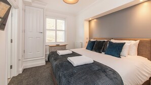 A bedroom with a large bed, a bedside table, a nightstand, a window with blinds, and a door.