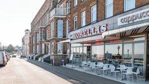 A street view with buildings, including a cafe named 'Morellis' and a 'Cappuccino' shop. There are outdoor tables and chairs, and a parked car on the side.