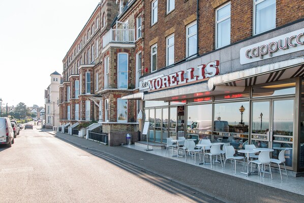 Vista de una calle con edificios, incluyendo un café llamado 'Morellis' y una tienda 'Cappuccino'. Hay mesas y sillas en la calle, y un coche aparcado al lado.