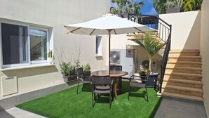 Een terras met een ronde tafel, stoelen en een witte parasol. Er staan potplanten en een trap die naar een hoger niveau leidt.