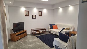 Een woonkamer met een televisie op een houten standaard, een bank met kussens, een salontafel en een vloerkleed.