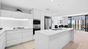 A modern kitchen with white cabinetry, a central island, and a view of a cityscape through large windows. - Dream Getaway on Level 29 with Breathtaking Ocean Views in Oracle Broadbeach! (Gold Coast)