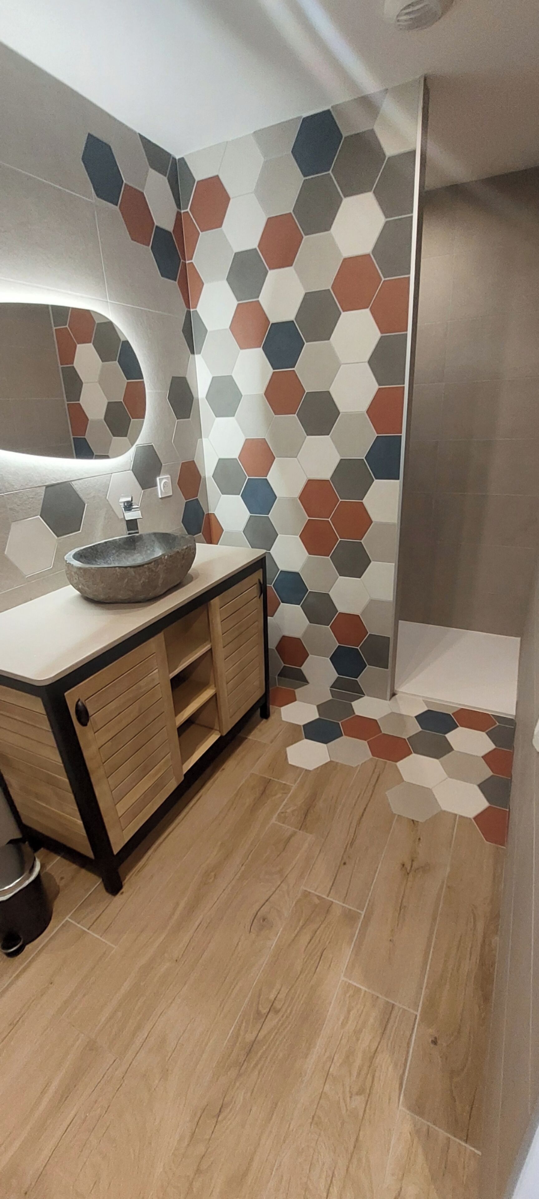 Een moderne badkamer met een houten wastafelmeubel, een rond wastafel en een spiegel. De wanden zijn betegeld met hexagontegels in verschillende kleuren.