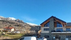 Un edificio moderno de dos pisos con fachada de piedra y ventanas grandes, situado en una zona montañosa con picos nevados al fondo.
