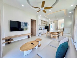 Una sala de estar moderna con un televisor de pantalla plana, una mesa de centro de madera, un sofá con cojines verde azulado, una zona de comedor con una mesa y sillas de madera, y una cocina al fondo.