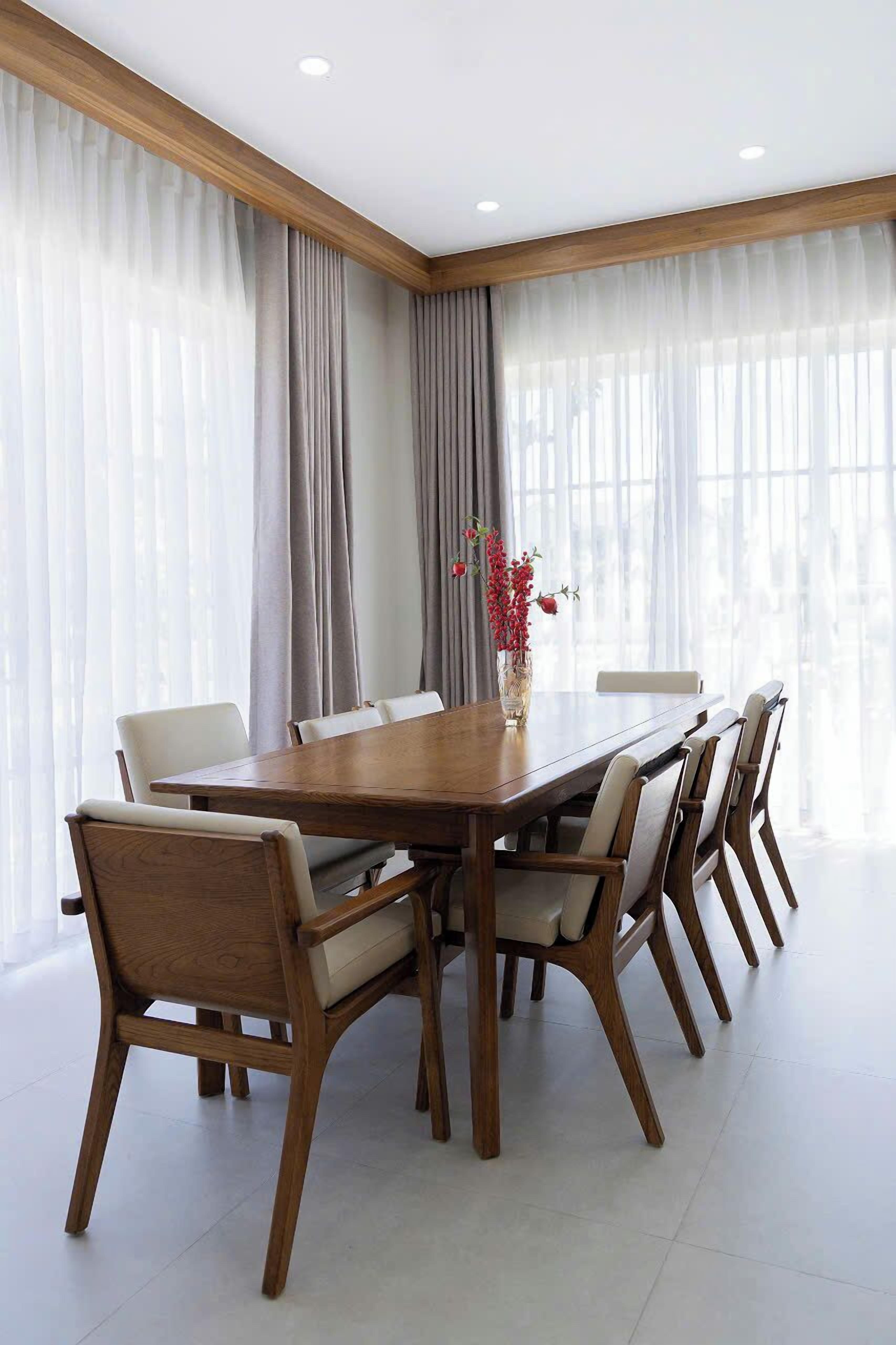 A conference room with a wooden table and chairs, large windows with sheer curtains, and a vase with red flowers.