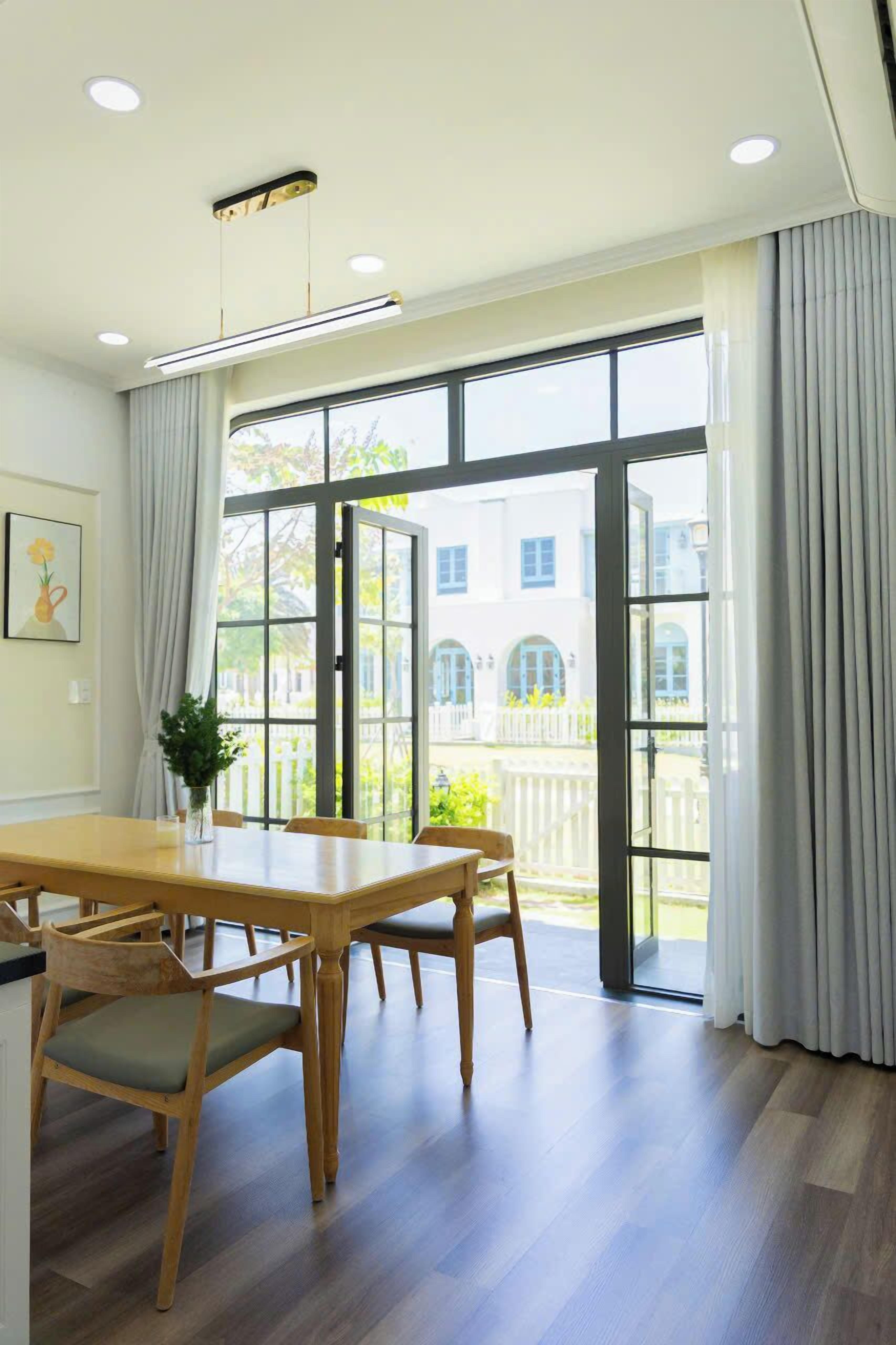 A dining area with a wooden table and chairs, large windows with curtains, and a view of a white building with arched windows.