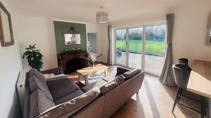 Een woonkamer met een bank, een open haard, een eettafel en uitzicht op een tuin door grote schuifdeuren.