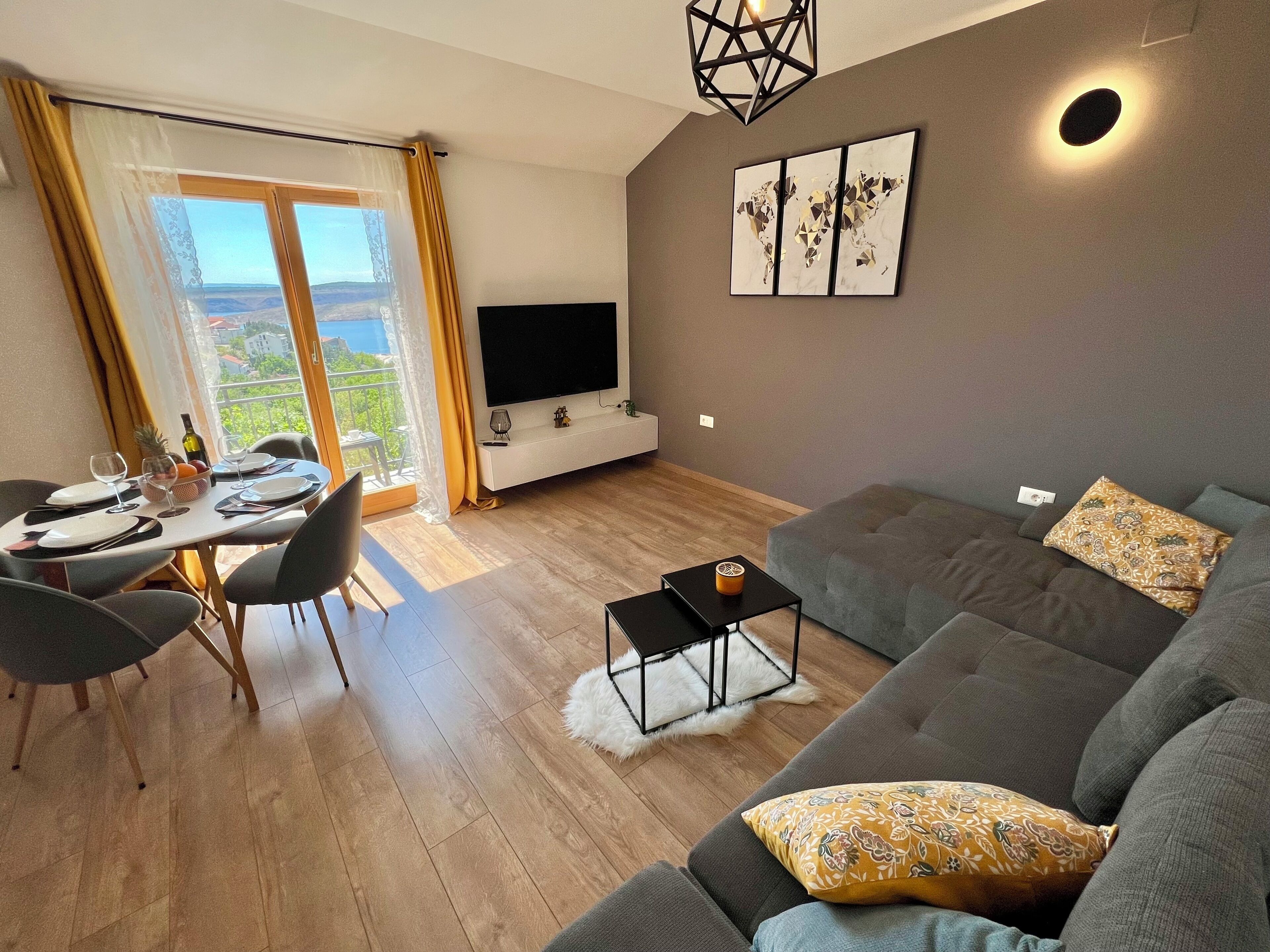 Een moderne woonkamer met een grijze bank, een eettafel gedekt voor twee personen, een flatscreen televisie en een groot raam met gordijnen.
