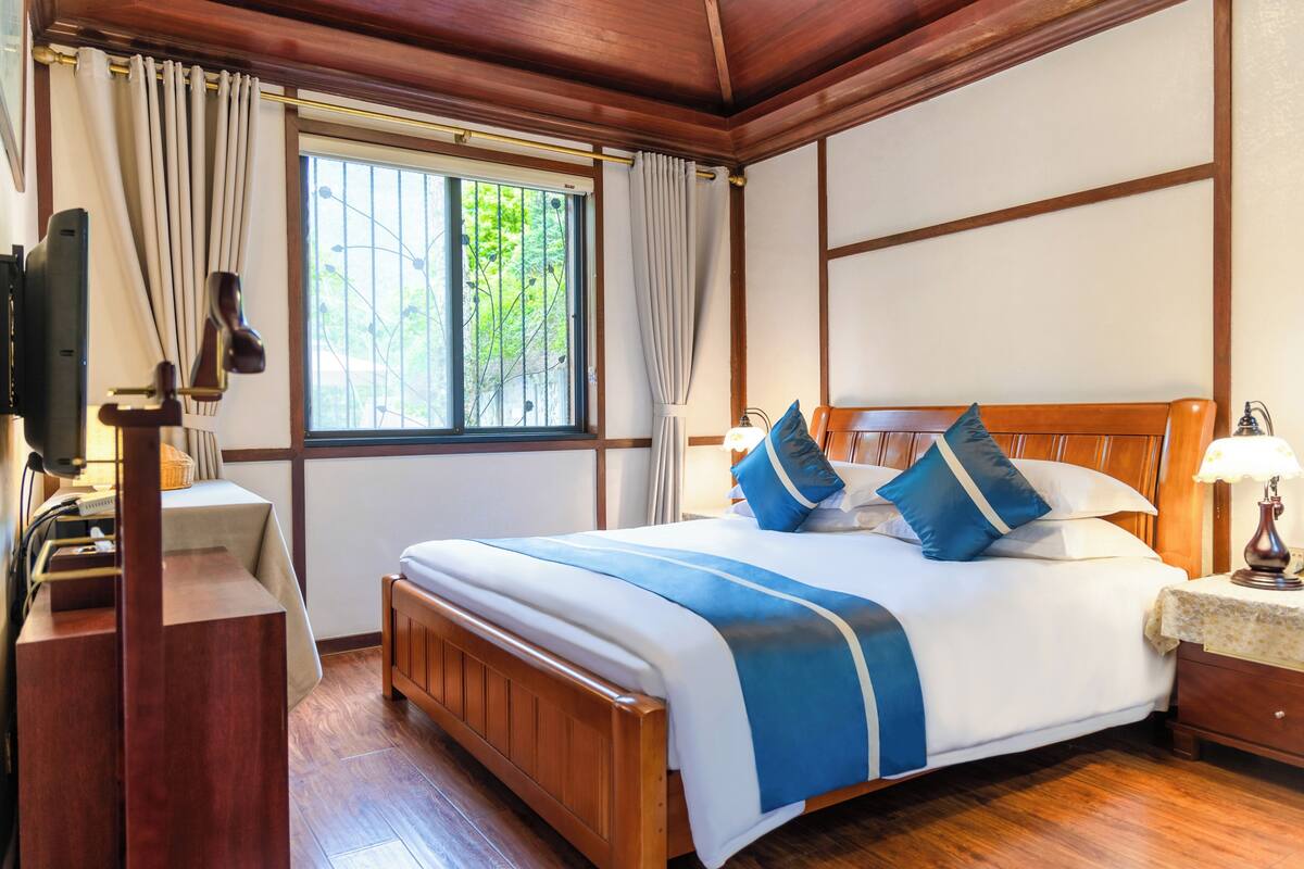 Une chambre d’hôtel bien rangée, avec un lit en bois, un bureau doté d’une lampe, une fenêtre avec des rideaux et un téléviseur posé sur un meuble.