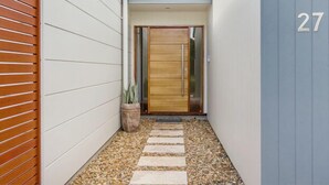 Une entrée moderne avec une porte en bois, le numéro « 27 » affiché sur le mur, et un chemin de gravier menant à la porte.