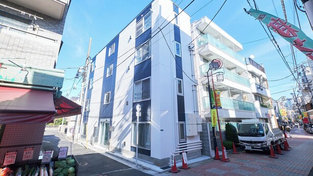 A multi-story building with a white and blue facade, a visible ground floor entrance, and a parked truck on the street. - Rio Grande Nishi-Shinjuku (Tokyo)