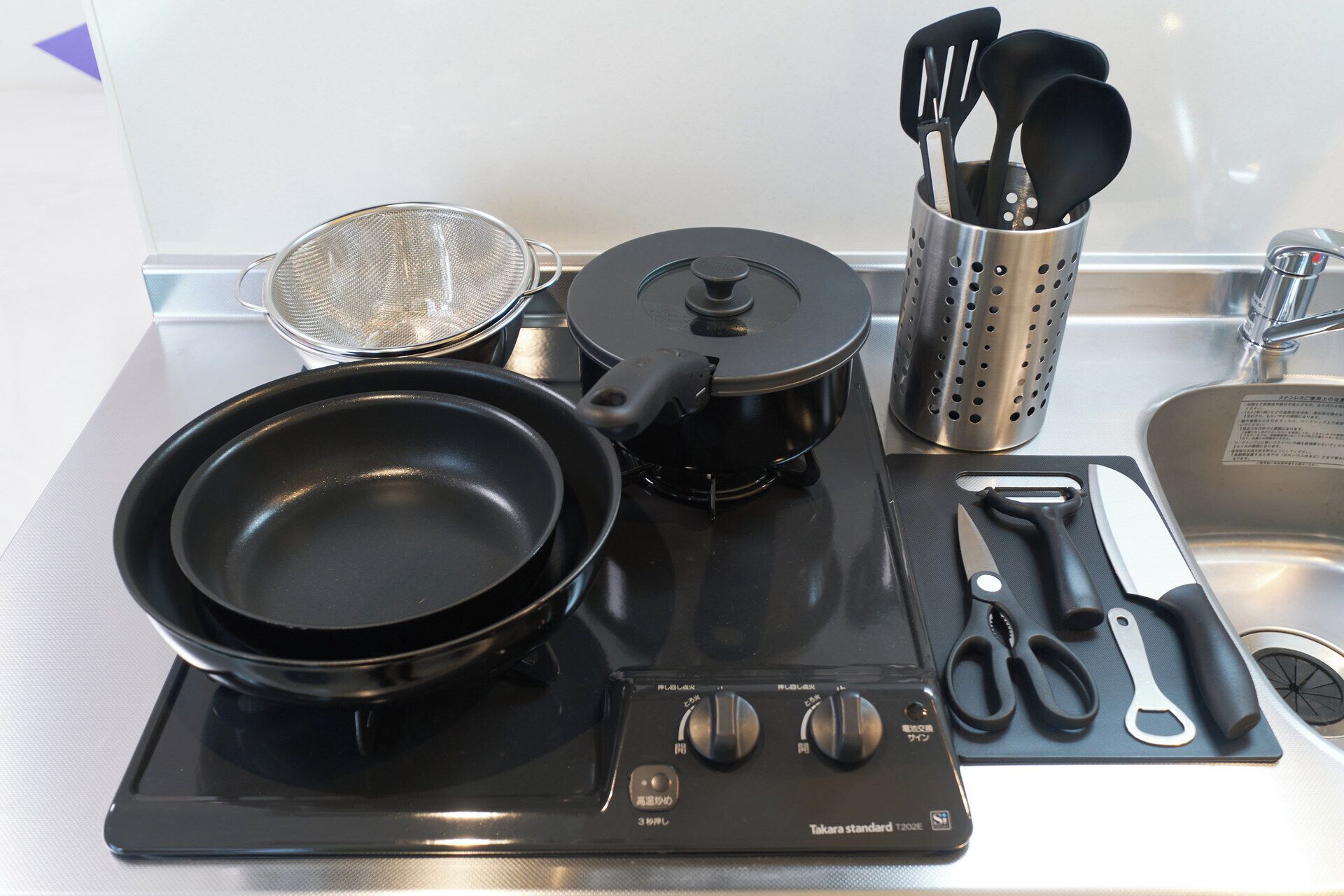 A kitchen with a Takara standard T2035 induction cooktop, a frying pan, a pot, a strainer, a utensil holder, and a cutting board with scissors and a knife.