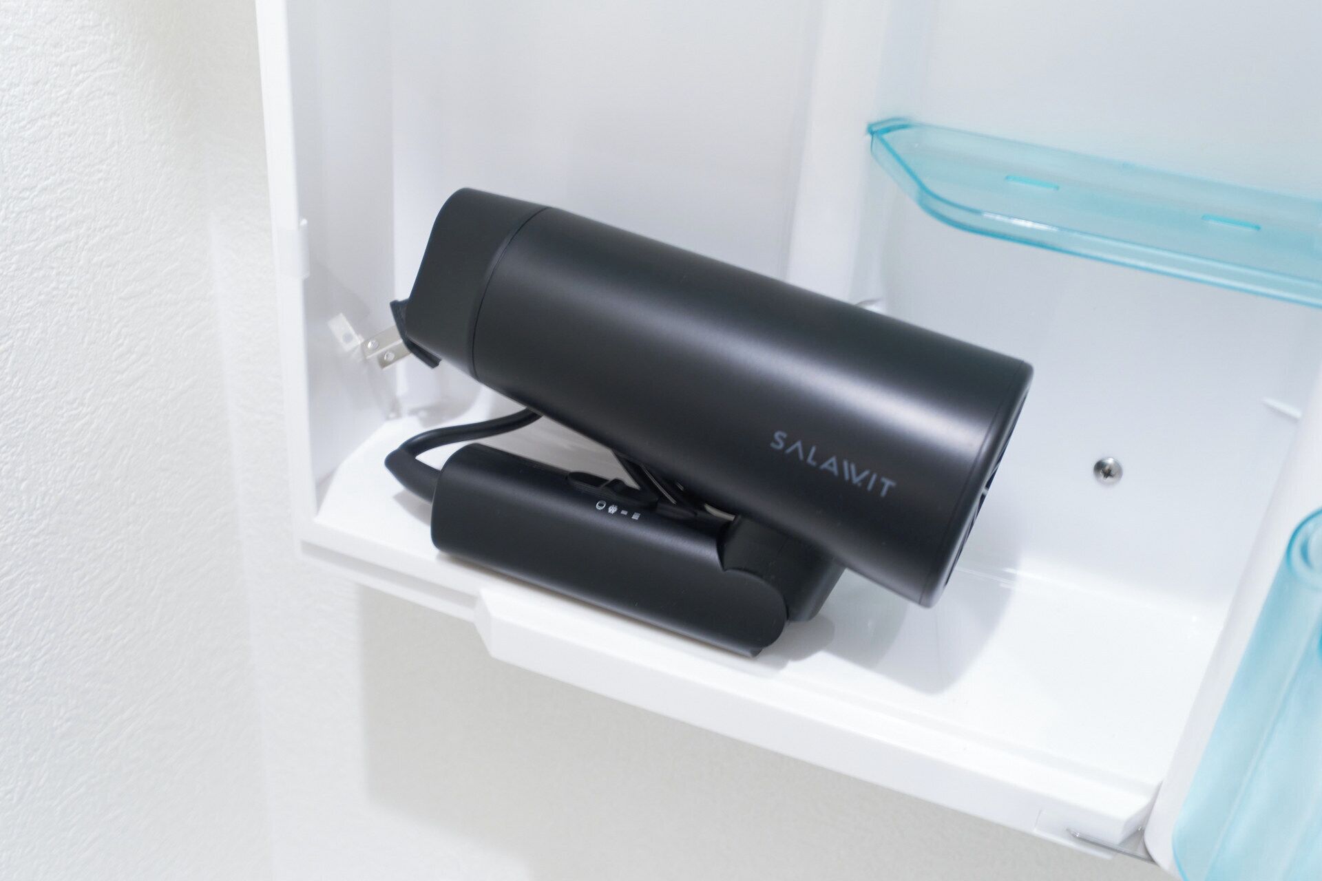 A black hair dryer and a blue container on a white shelf in a bathroom.