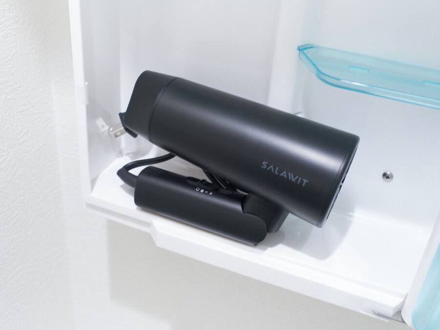 A black hair dryer and a blue container on a white shelf in a bathroom.