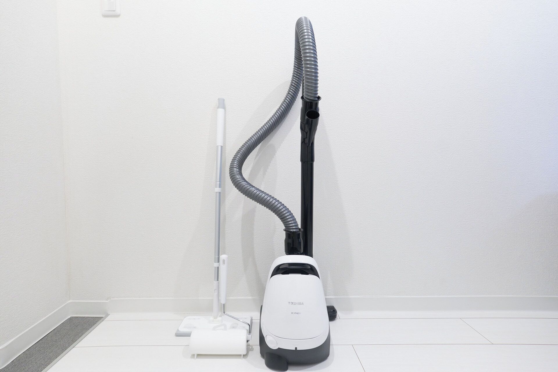 A white vacuum cleaner with a grey hose and a black power cord, placed on a white tiled floor against a white wall.