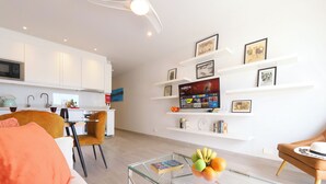 Un salon moderne avec une table basse en verre, un canapé et un coin repas avec des chaises. Des étagères blanches, ornées de tableaux encadrés, sont fixées au mur.